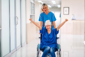 Pflegefachkraft in blauem Shirt schiebt fröhliche Patientin in blauem Shirt und Jeans im Krankenhaus zur nächsten Behandlung